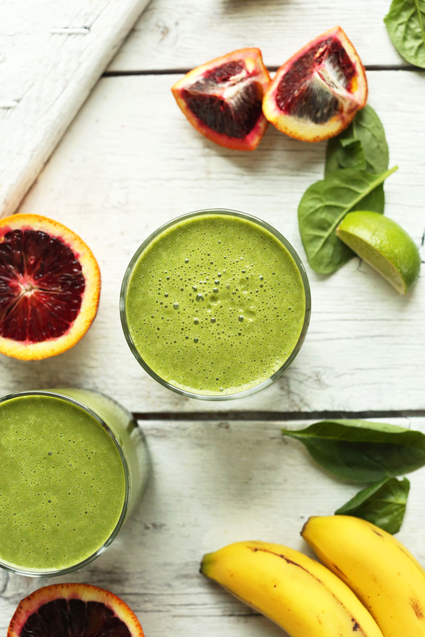 Two glasses of our Blood Orange Green Smoothie recipe beside fresh spinach, lime, blood oranges, and bananas